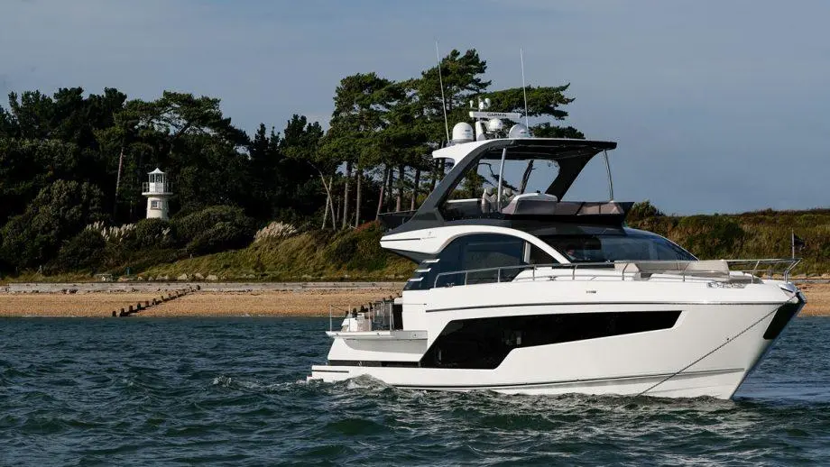 A sleek white motor yacht glides through calm waters near a sandy beach, with a lighthouse and trees in the background.